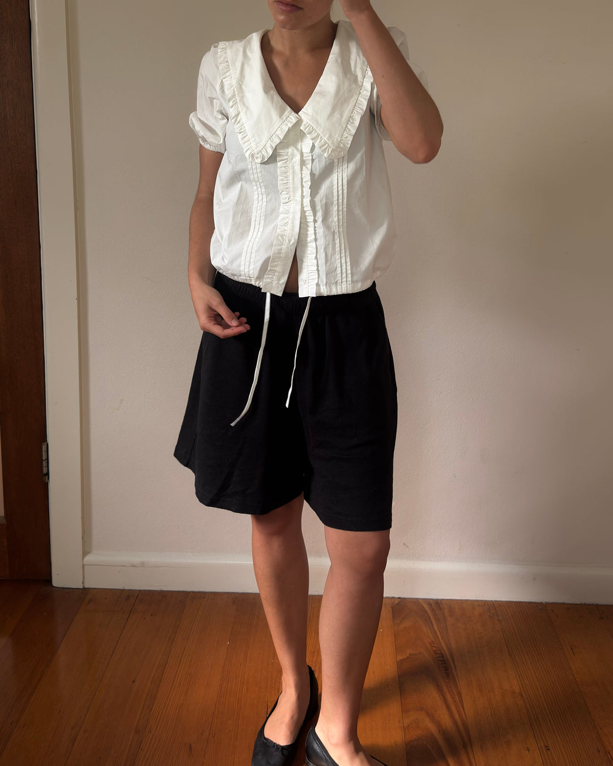 Person wearing a white blouse with ruffled collar and black shorts in a room with wooden floor and white wall.