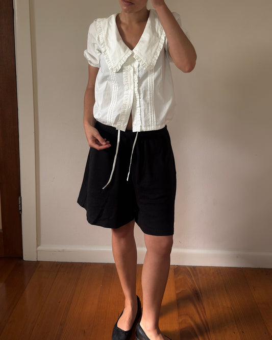 Person wearing a white blouse with ruffled collar and black shorts in a room with wooden floor and white wall.