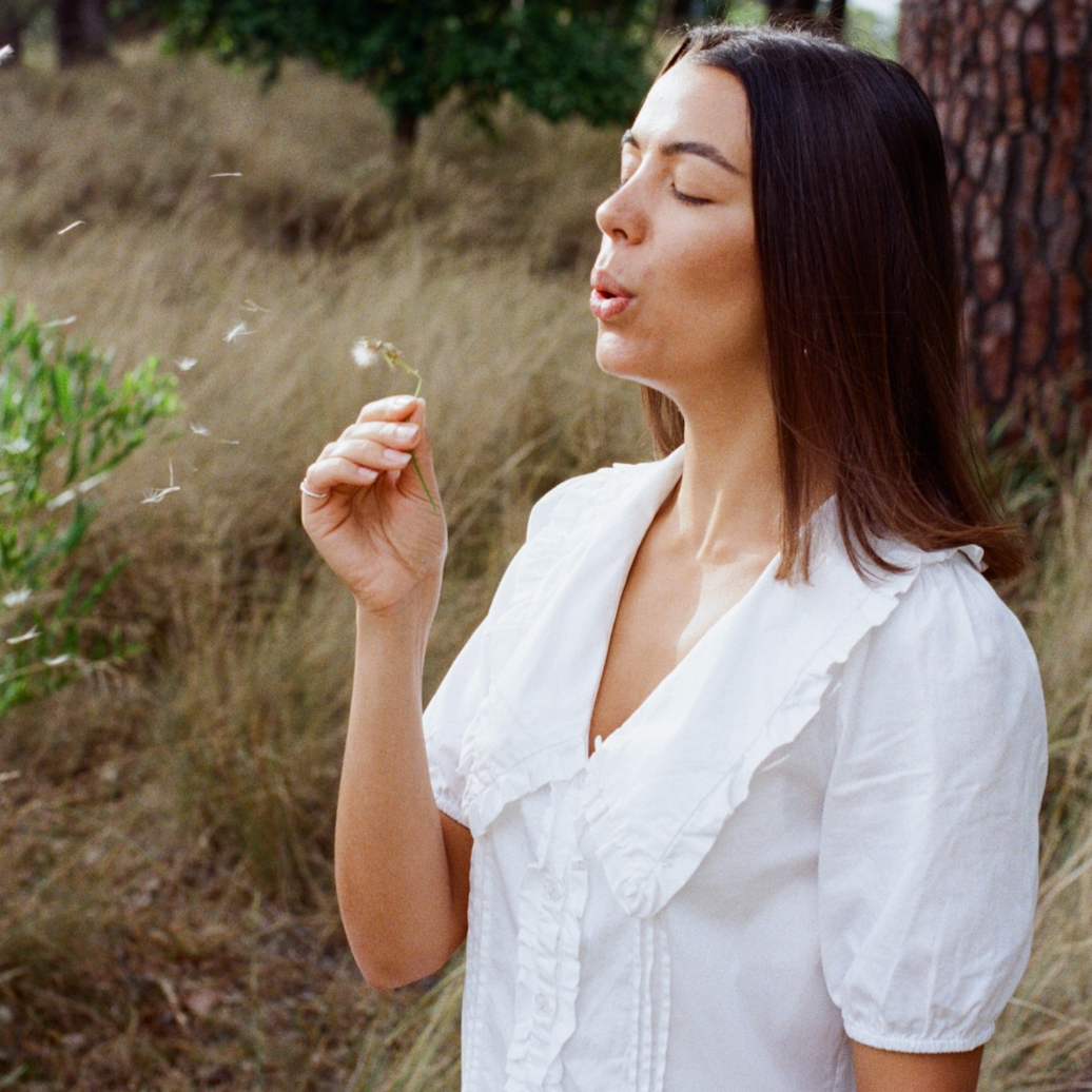 Woman blowing on a dandelion in a natural setting with trees and grass.